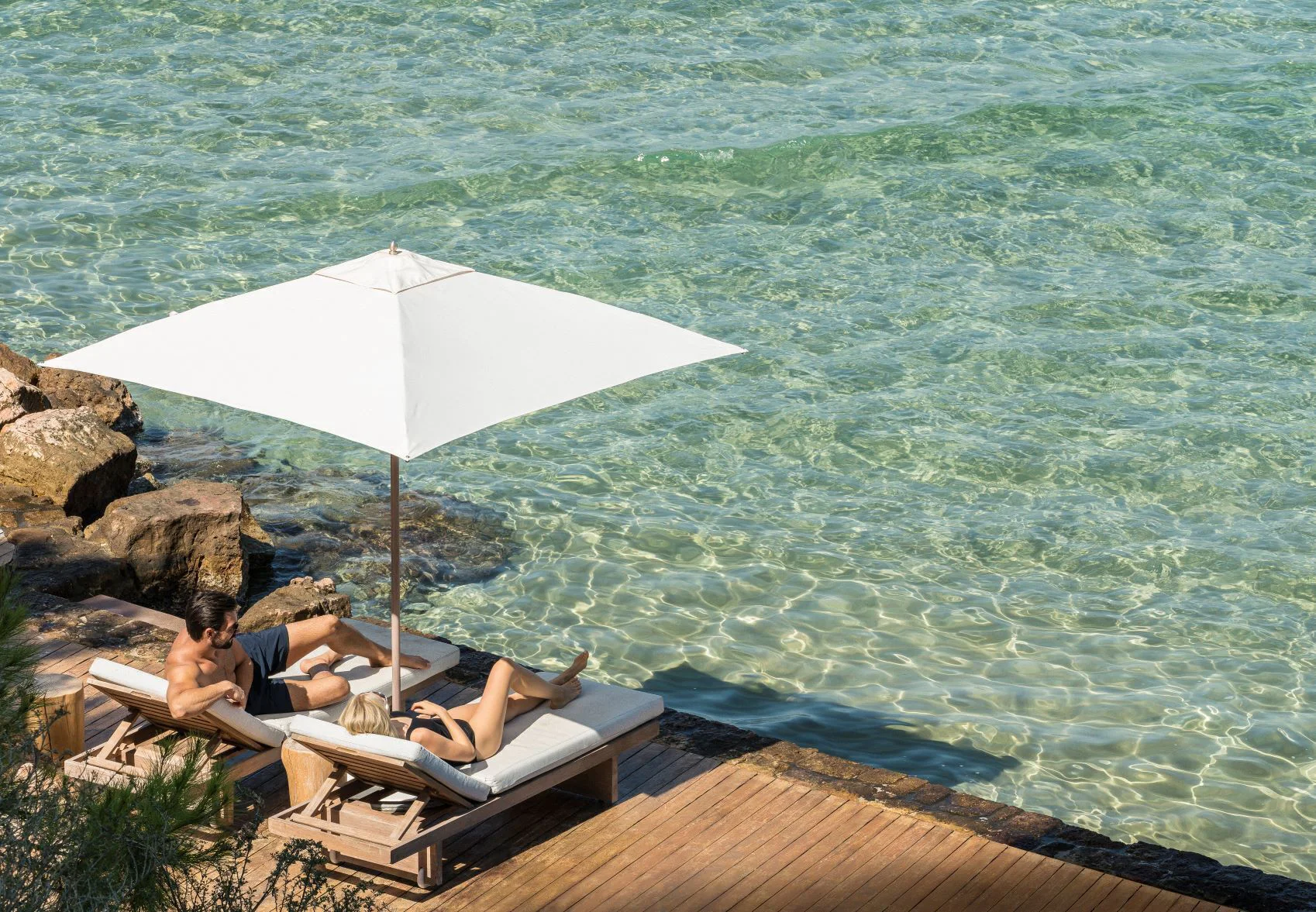 A couple relaxing on timber loungers beside the clear water of a private resort cove