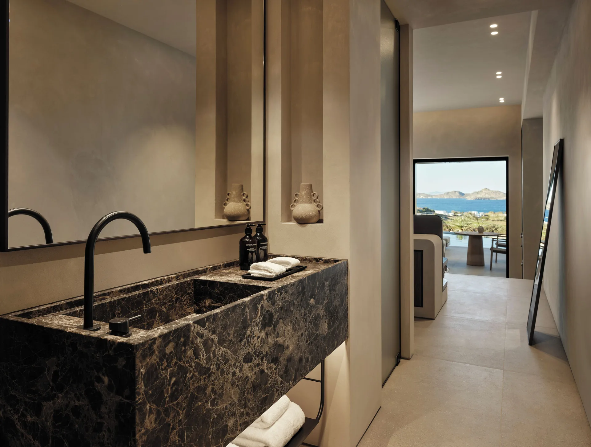 Dark marble suite vanity with a sightline through the bedroom to the blue Aegean